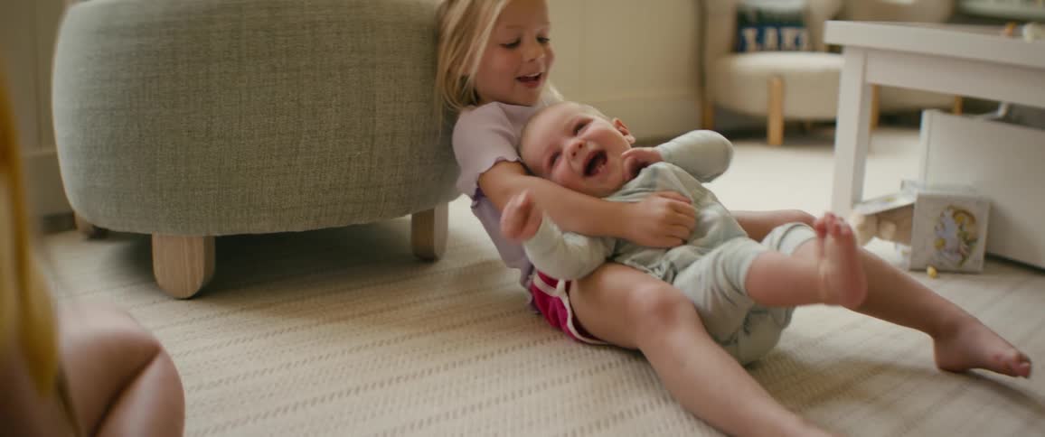 A mother tickles her laughing baby boy as he sits in his older sister's lap on the living room floor