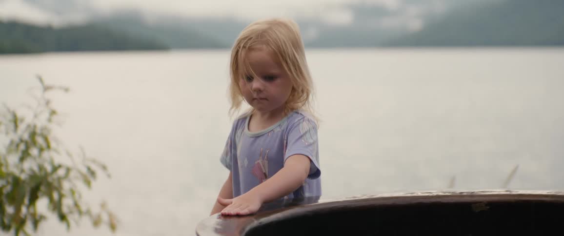 A thoughtful young blonde girl with a pensive expression rides in a boat across a calm lake on an overcast day