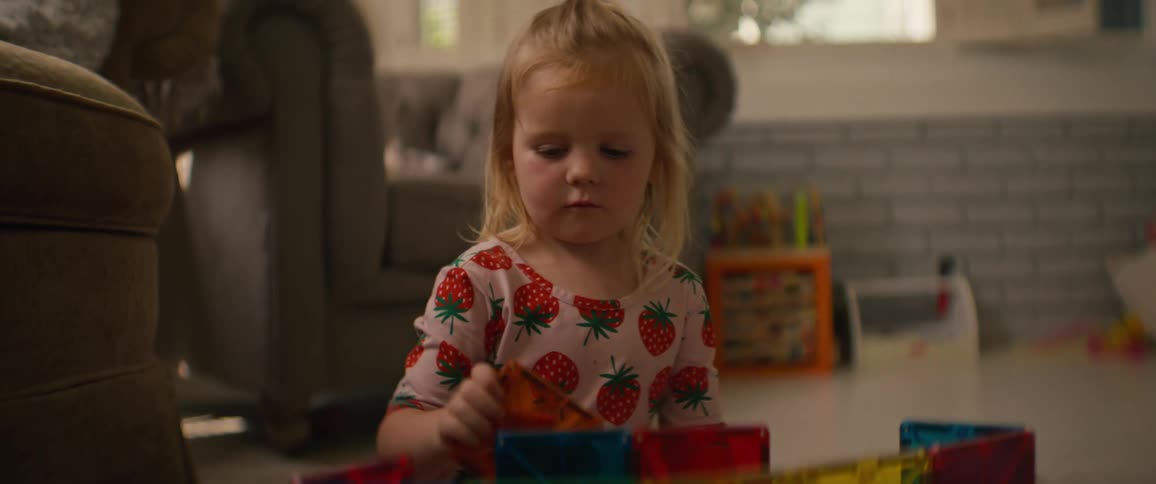 A cute toddler in a strawberry shirt intently plays with colorful magnetic tiles before rubbing her tired eyes