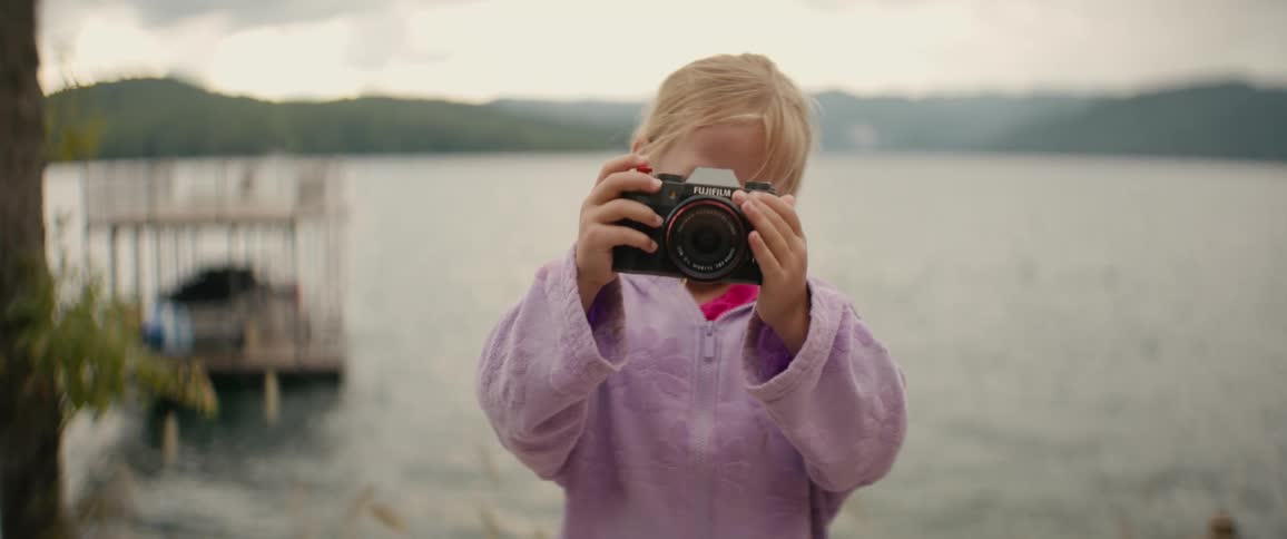 An adorable little girl with blonde hair takes a picture with a mirrorless camera while standing by a calm lake