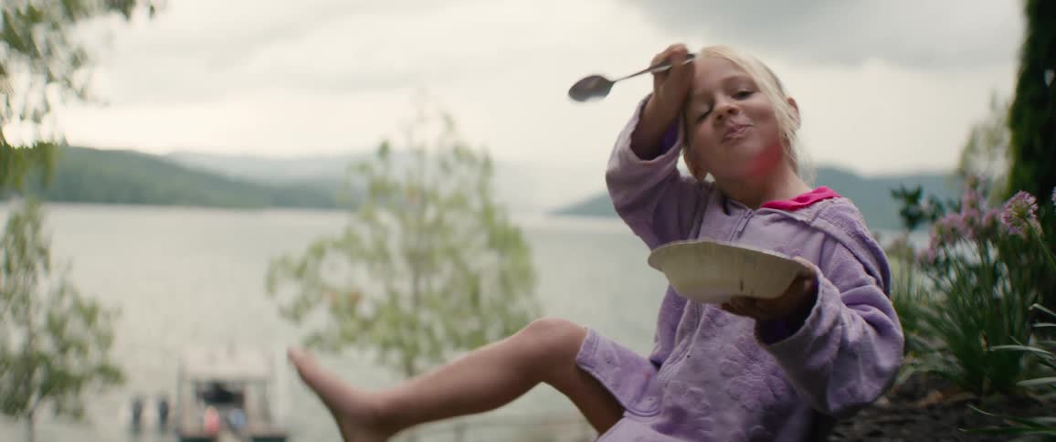 A happy young girl in a purple robe playfully eats a treat from a bowl by a lake
