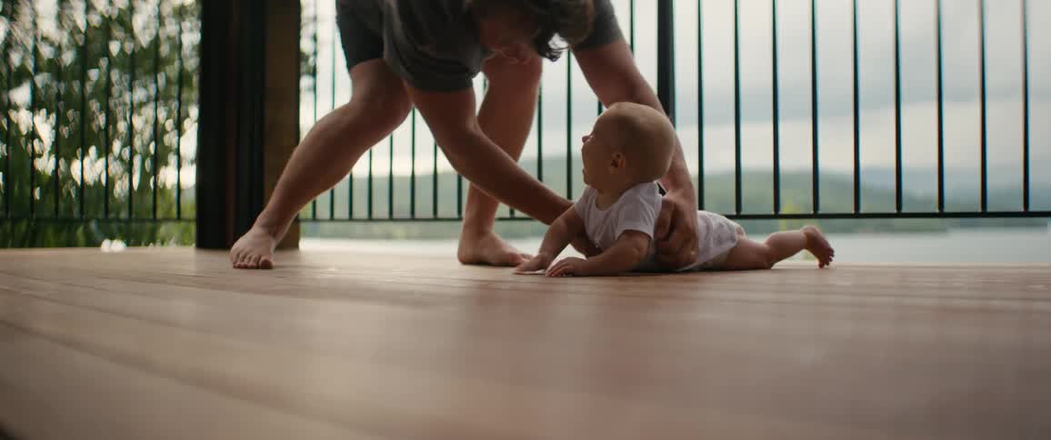 A father playfully tickles his baby during tummy time on a porch before picking her up and walking away, followed by a little girl running past