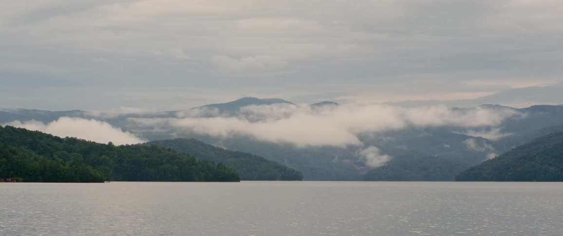 Misty clouds drift over the rolling hills and a calm lake on a gray, overcast day