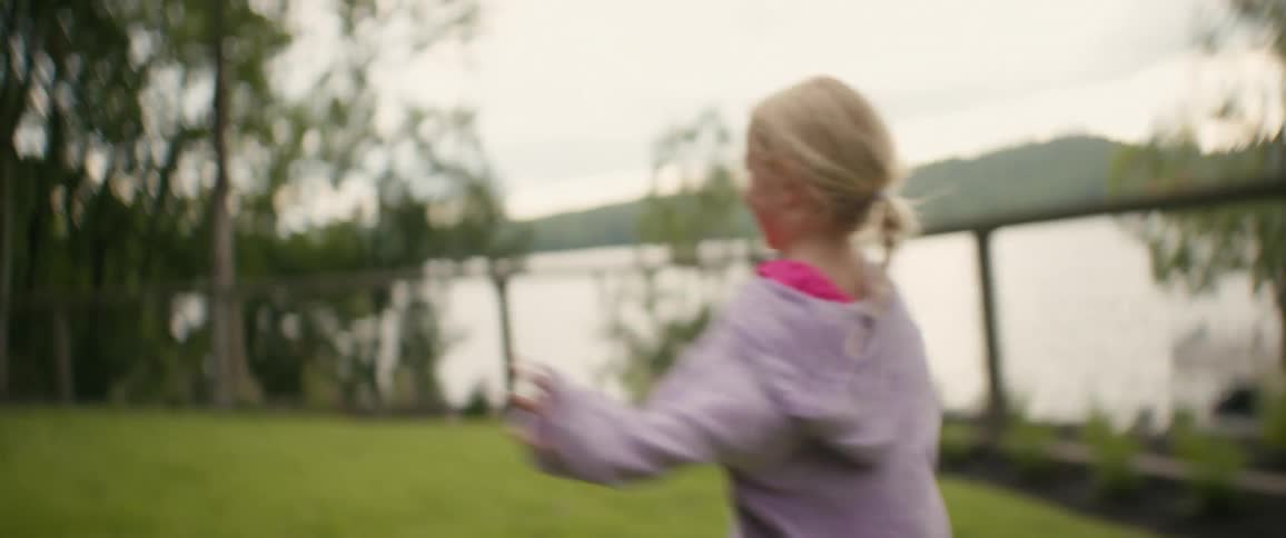 A happy little blonde girl smiles before running playfully across a lush green lawn in a beautiful slow-motion tracking shot