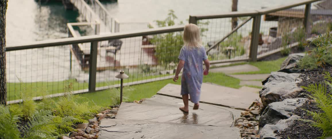 A little blonde girl in a purple dress walks barefoot down a stone path towards the water