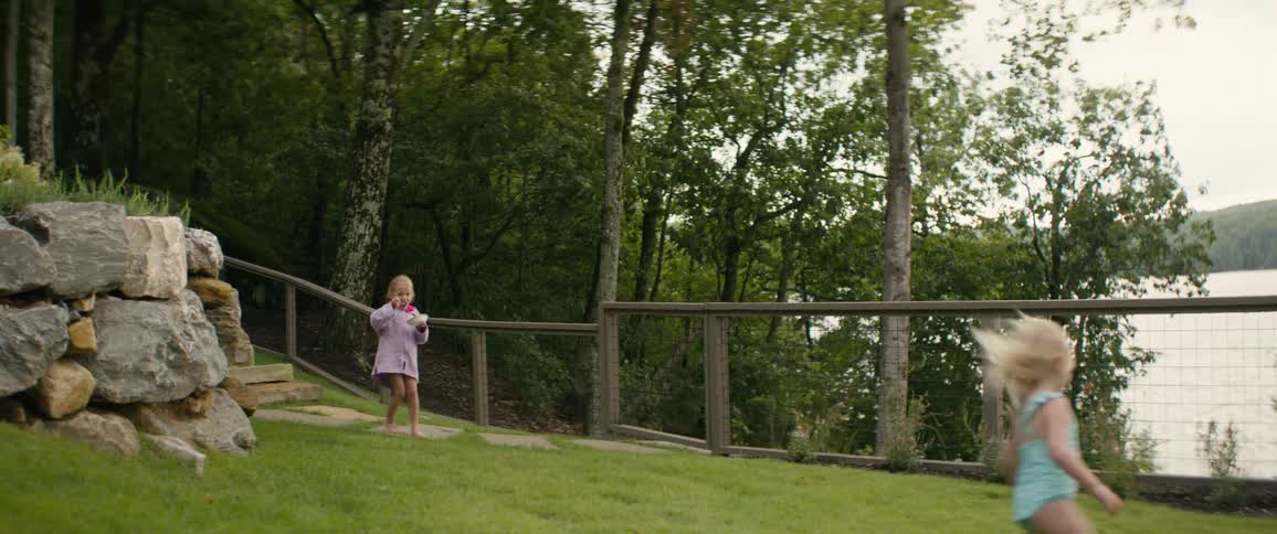 A young girl running through a green backyard while her sister watches from a distance
