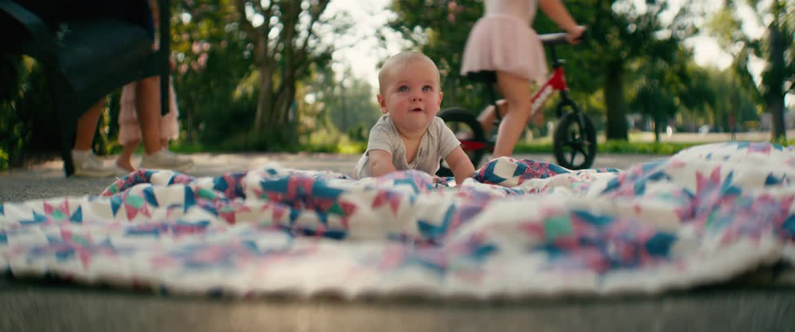 An adorable baby smiles on a blanket in the park while his older sisters play in the background