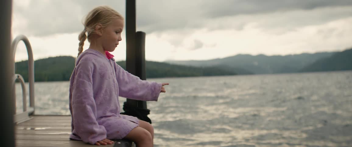 Little girl sits on a wooden dock and points across a calm mountain lake under dramatic cloudy skies