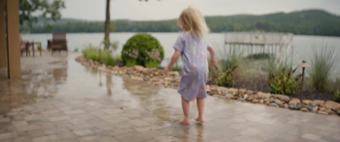 A young girl in a nightgown jumps barefoot on a wet stone patio with a lake in the background
