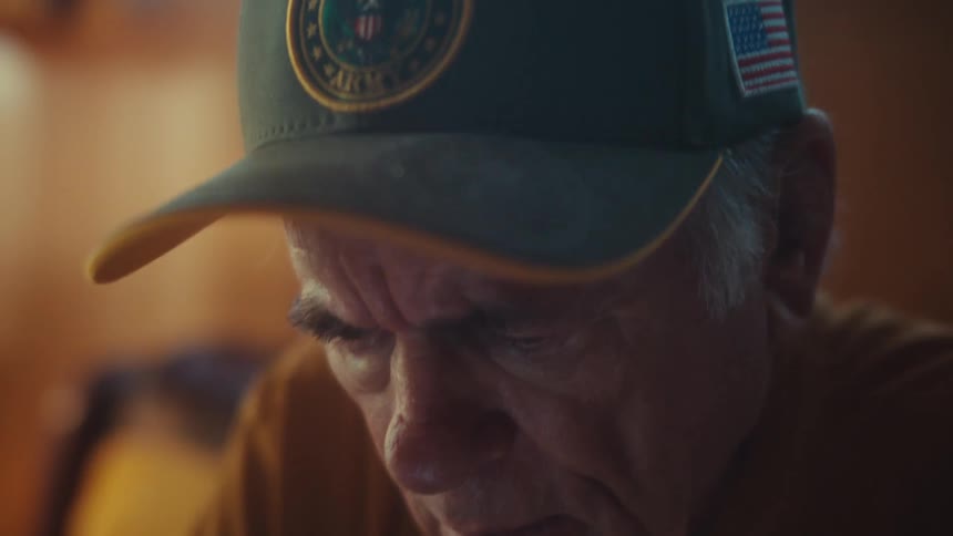 A solemn elderly U.S. Army Veteran sits thoughtfully as the camera tilts up to reveal his hat