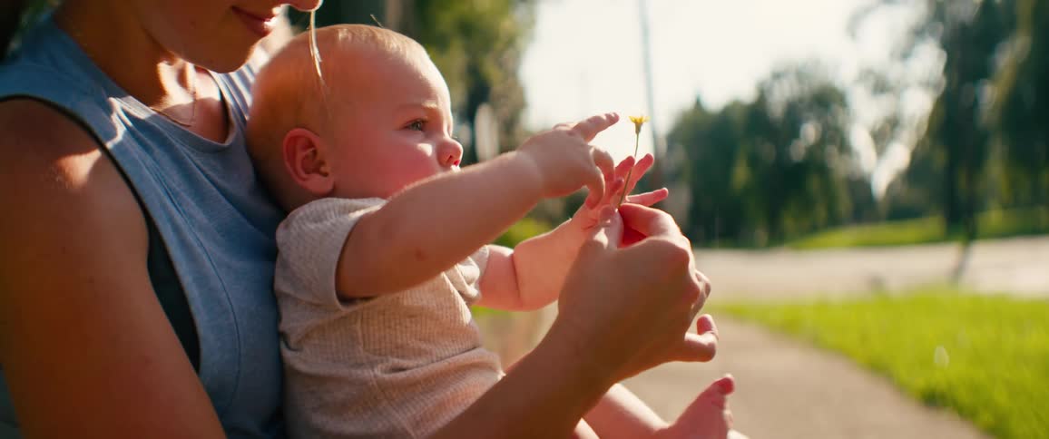 A mother holds a small yellow flower for her curious baby to touch and explore while sitting in her lap on a sunny day