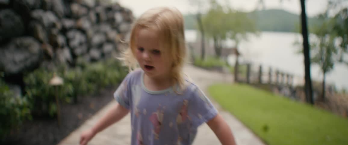 An adorable blonde toddler in pajamas walks down a stone path towards the camera with a lake in the background