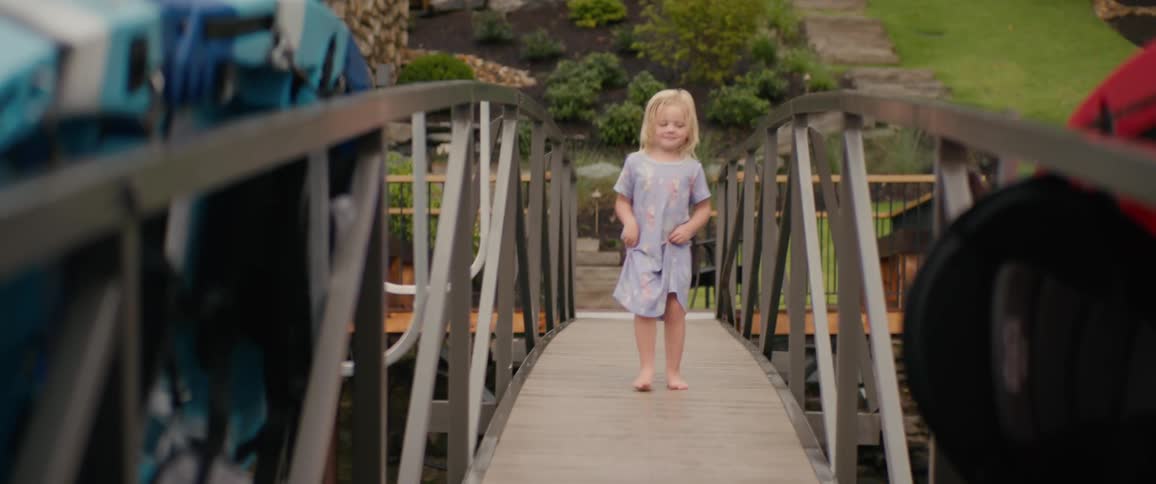 A cute barefoot toddler with blonde hair playfully walks and makes silly faces while walking on a ramp leading to a dock