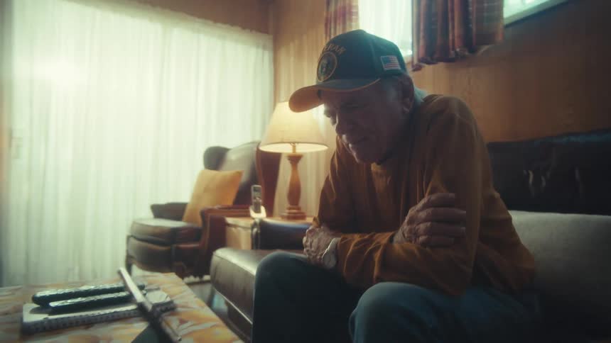 A sad elderly veteran sits alone in his dimly lit, vintage-style living room, looking down at a tablet
