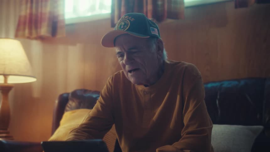 An elderly veteran wearing a cap laughs with joy while watching a video on a tablet in his cozy, lamp-lit living room, his expression becoming nostalgic