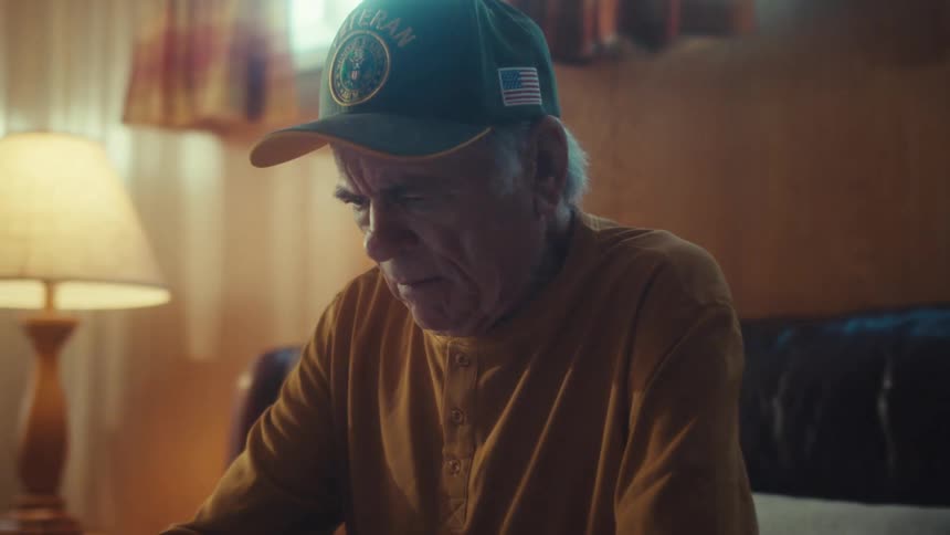 A pensive elderly veteran sits alone in his dimly lit living room, looking down at a tablet on his lap