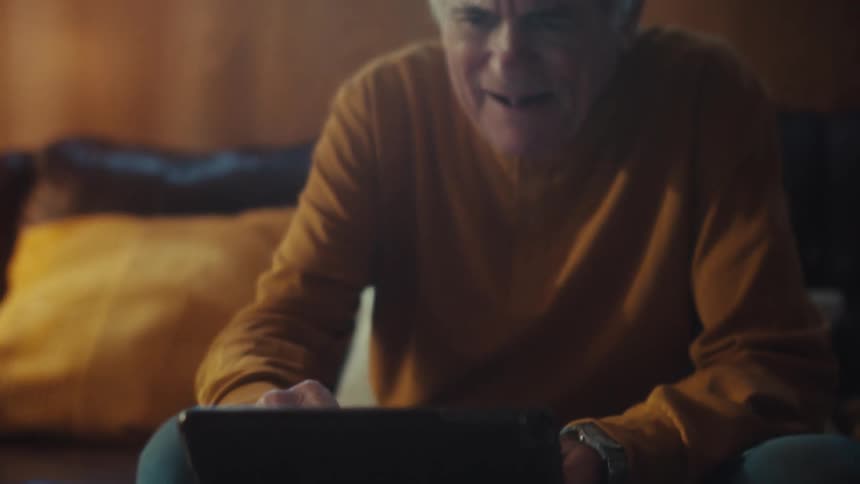 An elderly veteran in a cap smiles and laughs while watching something on his tablet in a warmly lit living room