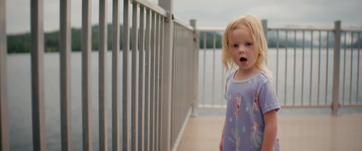 A close-up of a sweet blonde toddler looking up with a shy smile while standing on a pier overlooking a lake