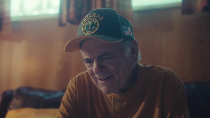 Elderly veteran in a cap smiles with joy while watching something on his digital tablet in a warm, cozy living room