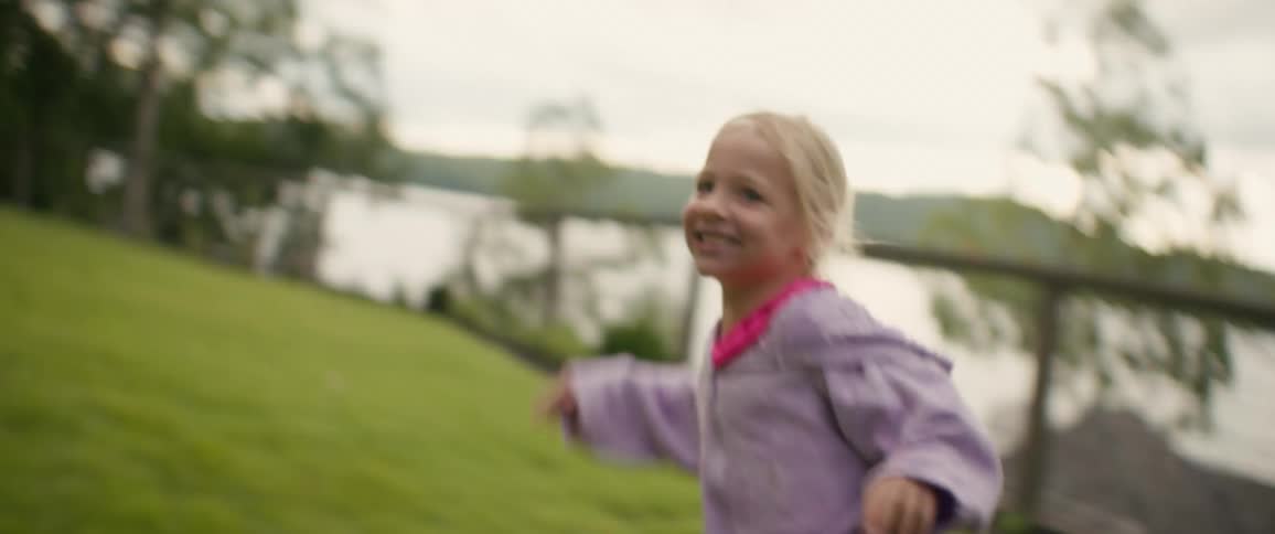 A joyful little girl with blonde pigtails runs and laughs in a grassy yard by a lake
