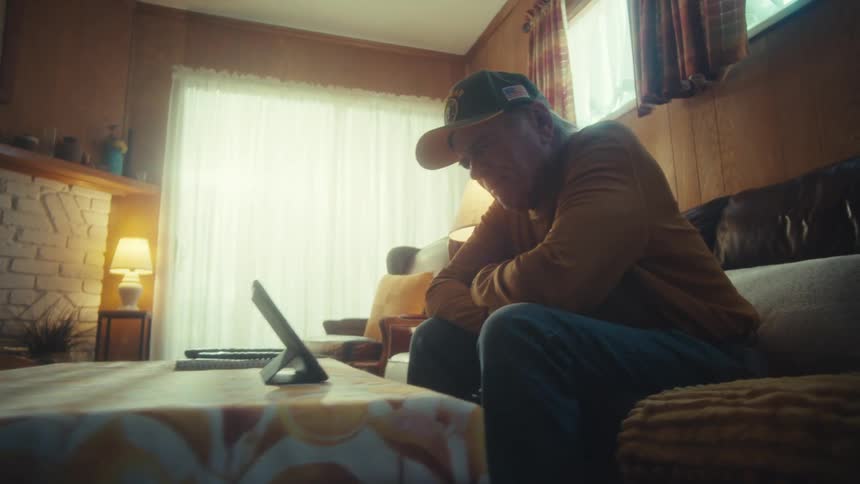 A lonely senior man sits on his couch in a vintage-style living room while watching something on a tablet