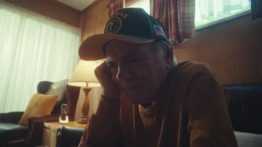 A lonely and pensive elderly veteran sits on a couch in his modest living room, looking thoughtfully at a tablet