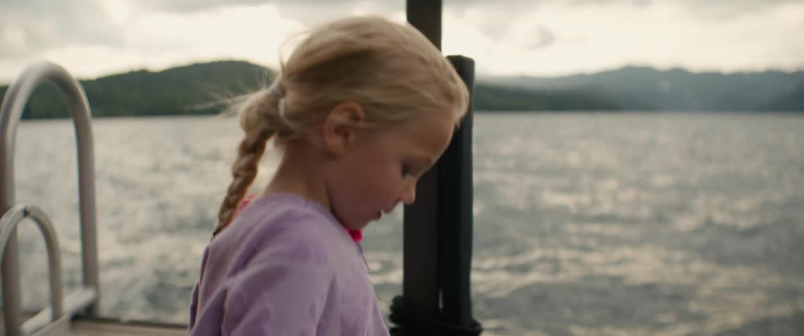 A young girl with blonde pigtails contemplates quietly on a boat as it moves across a tranquil lake