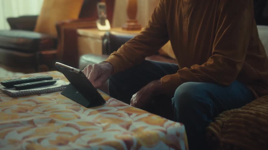An elderly veteran sits on a sofa in his living room, scrolling through content on his digital tablet