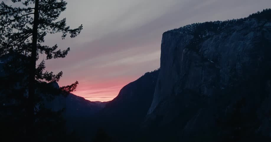 A majestic mountain cliff stands silhouetted against a beautiful pink and purple sunset over a deep valley