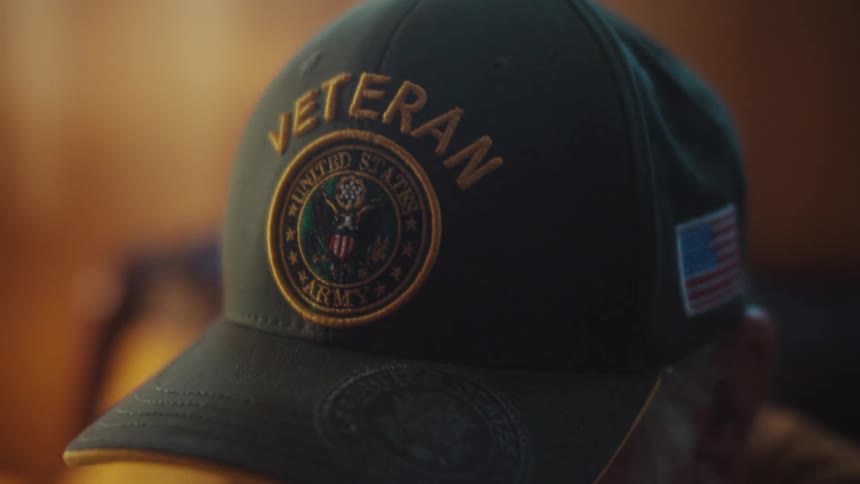 A close-up of a pensive elderly man wearing a United States Army veteran baseball cap