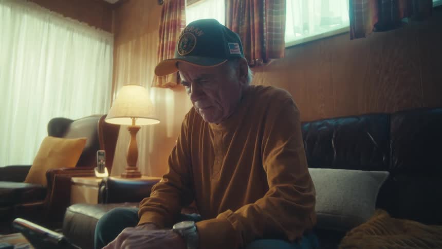 A slow zoom reveals an elderly veteran sitting alone on his couch, deeply engaged with his tablet in a softly lit room