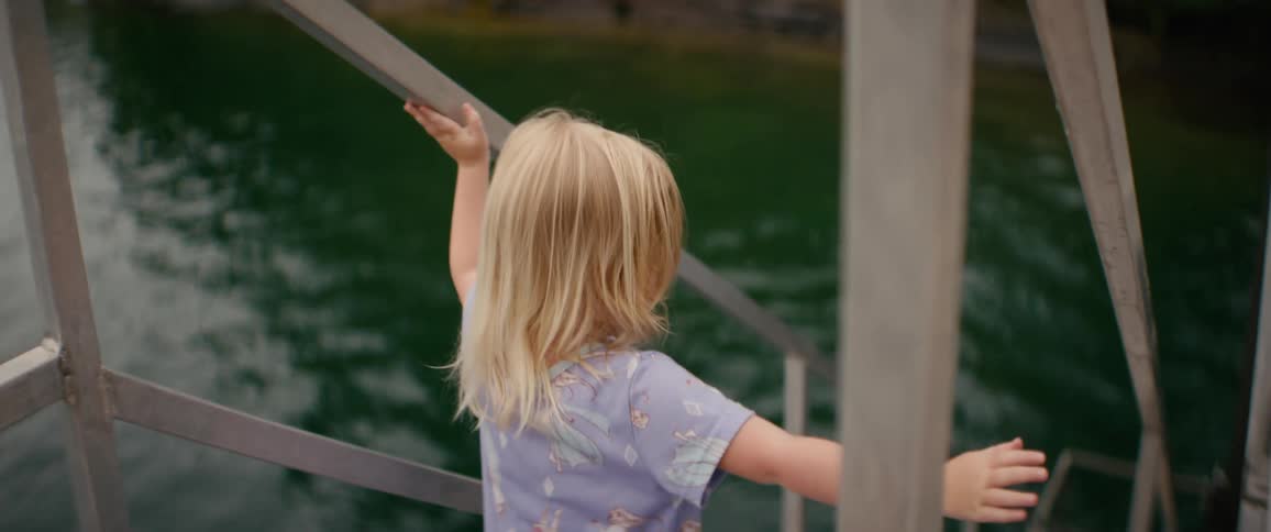 Following a curious blonde toddler in pajamas as she walks along a lakeside pier and down to the dock