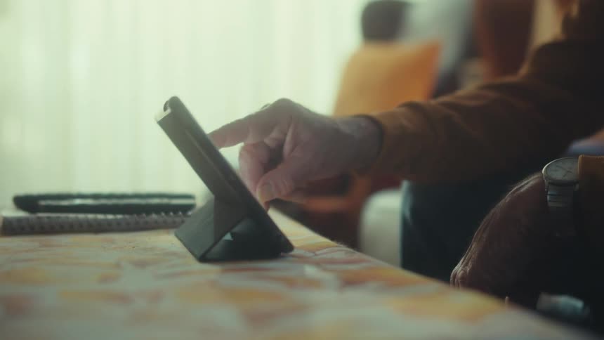 A senior veteran wearing a patriotic hat thoughtfully uses a tablet in his warmly lit home