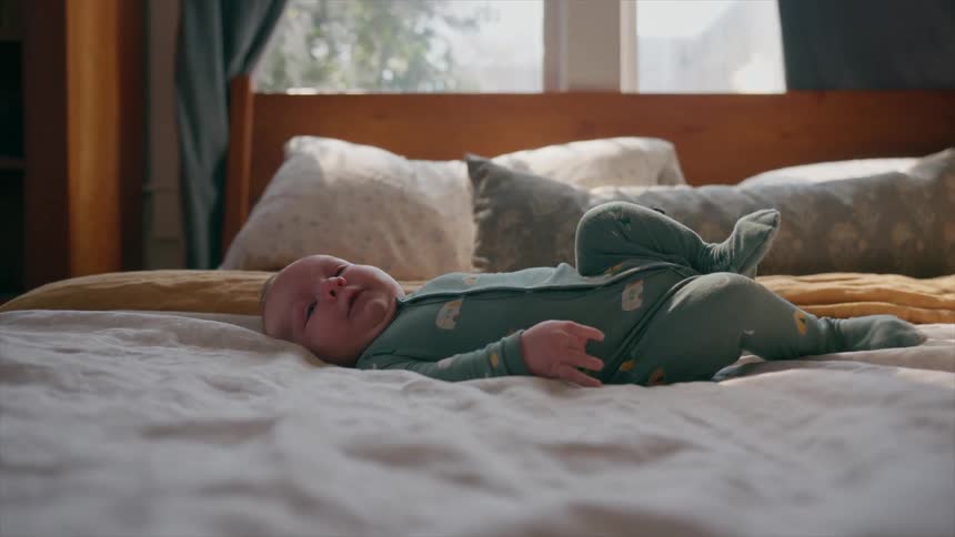 A calm newborn baby in a green sleeper lies on its back on a comfortable bed in a sunlit room