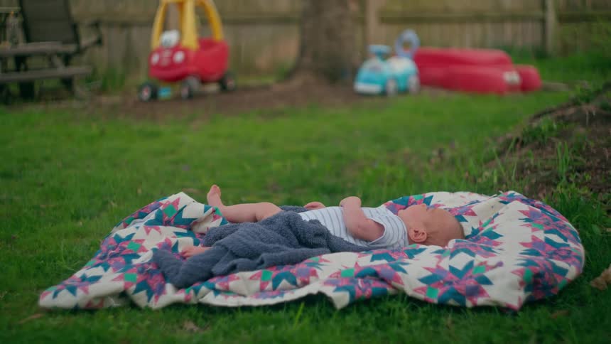 A calm baby boy lies on his back on a patterned quilt in the backyard with toys in the background