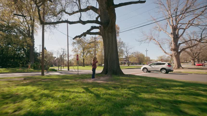 A cheerful little girl stands by a large tree in a sunny neighborhood park before smiling and running toward the camera
