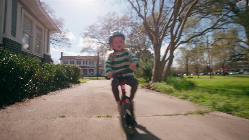 A happy little girl in a helmet laughs as she rides her red balance bike down a neighborhood sidewalk