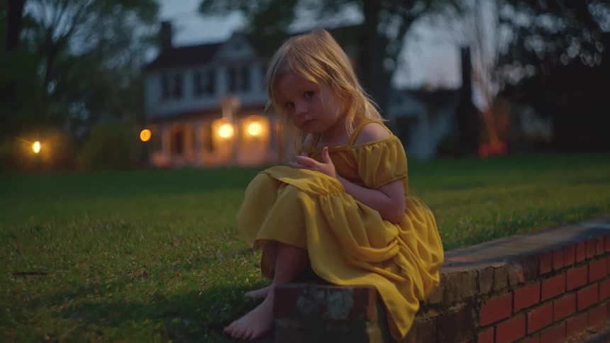 A barefoot toddler in a yellow dress sits sadly on a brick wall in her front yard at dusk