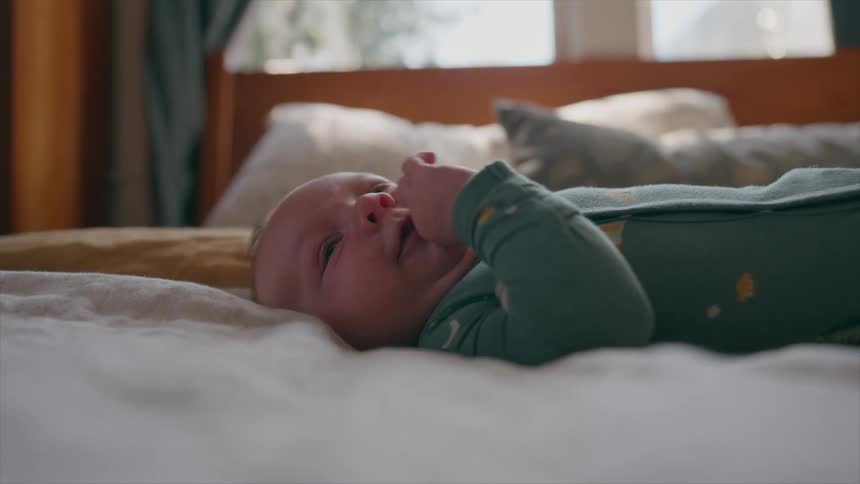 A peaceful newborn baby in a green onesie lies on a bed, looking around calmly and sucking on their fist