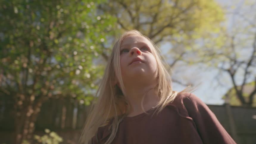 A young blonde girl stands in her lush backyard garden, looking around with wonder on a bright sunny day