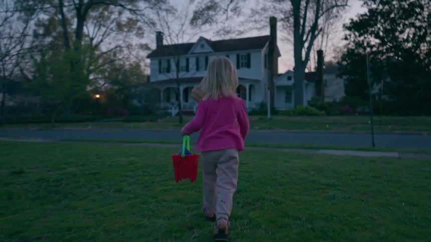 A little girl in a pink shirt runs across a green lawn with her red bucket to join her sister for an Easter egg hunt at dusk