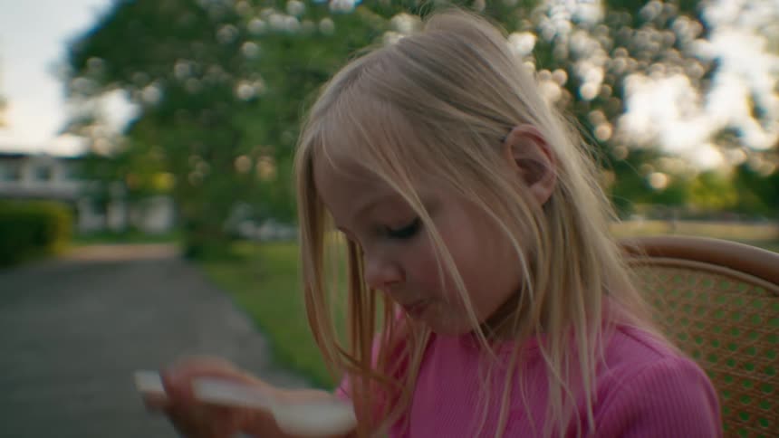 A cute little blonde girl in a pink shirt enthusiastically eats a spoonful of food outdoors at dusk