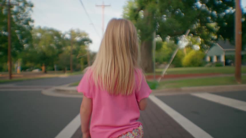 Rear view of a little blonde girl with long hair walking down a brick crosswalk in a quiet suburban town