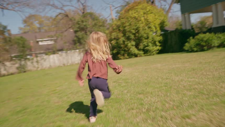 A carefree young girl with blonde hair runs across a sunny green lawn in a suburban park
