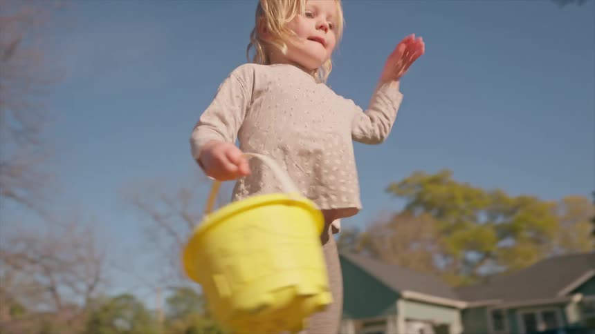 A barefoot toddler with a yellow bucket happily walks and balances on a brick wall, then tumbles off on a sunny day