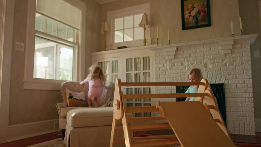 Two energetic sisters play together in a sunlit living room, climbing on a wooden playset and jumping on the furniture