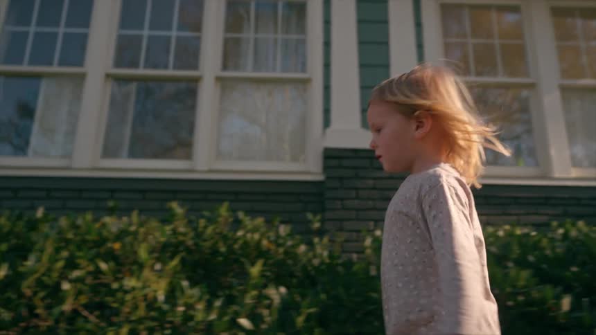 A little girl with blonde hair blowing in the wind walks with a yellow bucketi n front of a suburban home