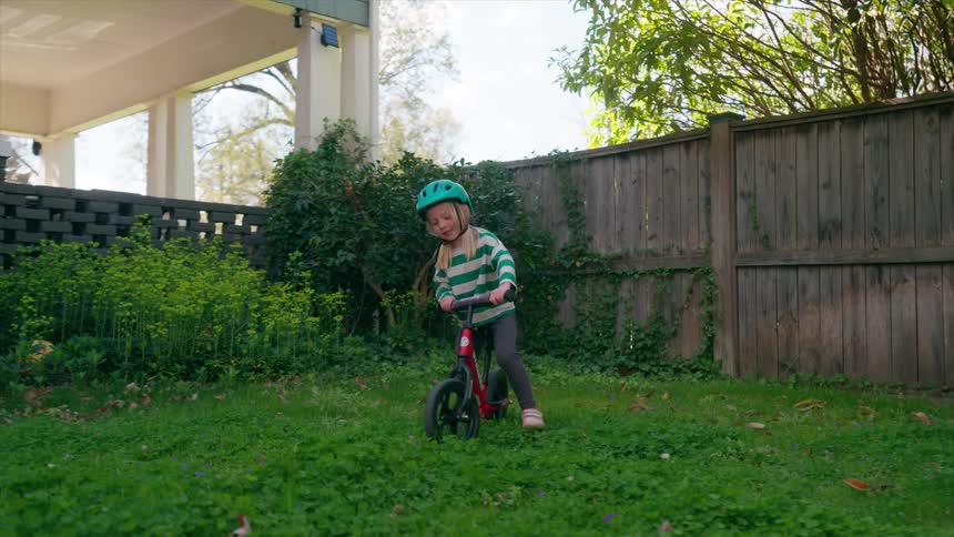 A determined little girl in a green helmet happily rides her red balance bike across the green grass in her backyard