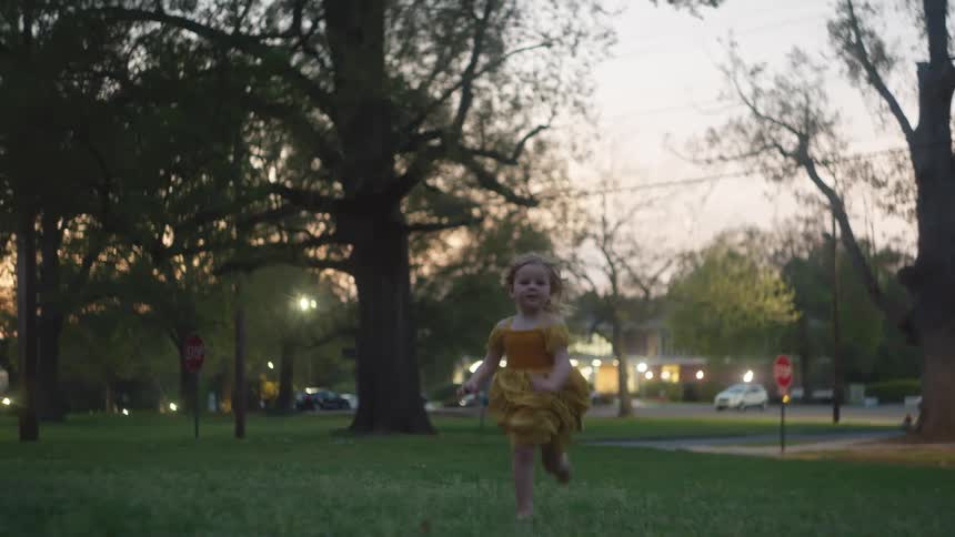 A joyful little girl in a yellow dress runs barefoot through the grass towards the camera at dusk