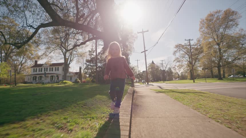 A happy little girl carefully balances as she walks along a low brick wall next to a sidewalk in a quiet, sunny neighborhood
